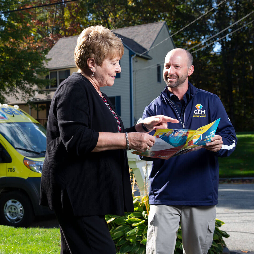 A GEM technician is showing a brochure to a female customer in her yard. They are standing beside a colorful GEM service van parked on the street. The background features a sunny suburban setting with a home and trees.